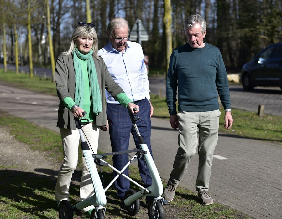 An older woman walking with a rollator alongside two companions